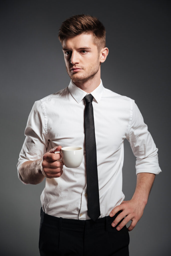 Successful young businessman in formalwear holding cup of coffee while standing isolated over grey background